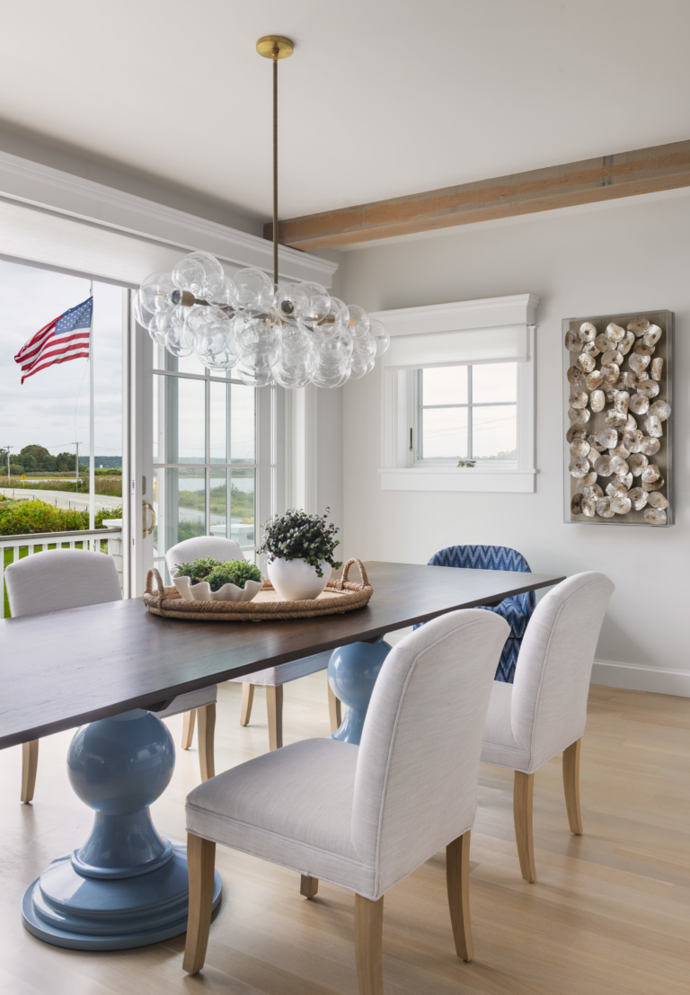 The interior design of a coastal-inspired dining room with an ocean view, chandelier, and exposed beams.
