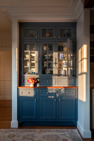 Custom blue built-in cabinetry with glass-front doors and curated ceramics, lit by late afternoon sun in the kitchen of Ocean Lodge.